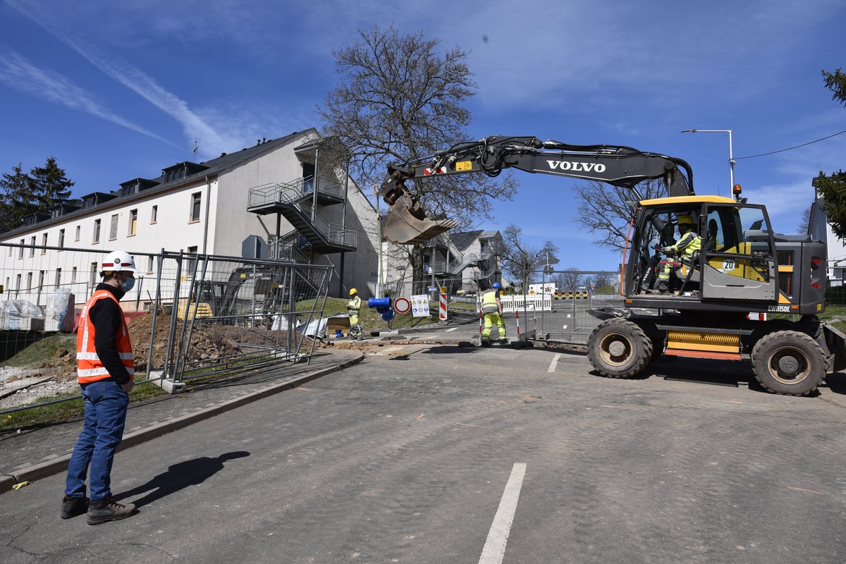 Excavator and crew working beside a barracks building under renovation, with a construction manager in a hard hat watching from the road.
